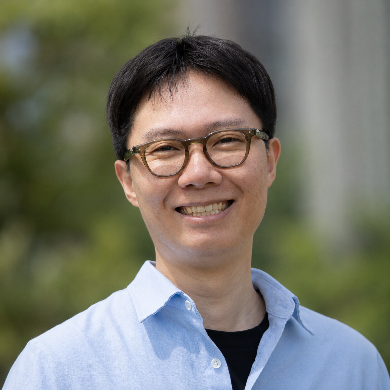 Photograph of a Asian man with short brown hair, eye glasses, and an blue shirt, standing in front of buildings and trees.